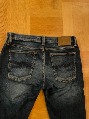 Mörkblå jeans från Nudie Jeans - Snygga mörkblå jeans från Nudie Jeans med klassisk femficksdesign och orangea kontrastsömmar. Jeansen har en rak passform och är tillverkade i slitstark bomull. Bakfickorna har Nudie Jeans typiska broderi och läderpatch med logga. OBS det står att dom är L34 men dom är uppsydda som ni ser på sista bilden