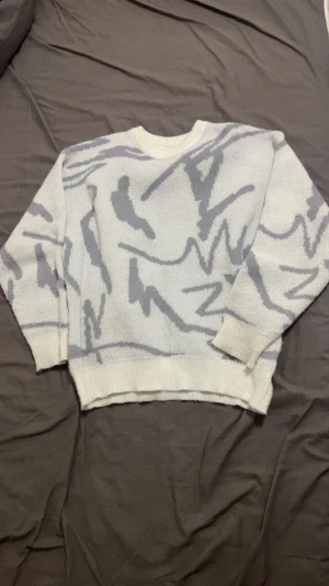 Vit och grå stickad tröja med mönster - Säljer en vit stickad tröja med grått abstrakt mönster. Tröjan har rund hals och ribbade muddar vid ärmslut och nederkant. Perfekt för dig som gillar unika prints och vill ha en mysig, stilren look.