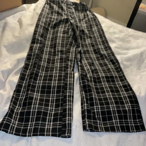 Svarta rutiga byxor från H&M - Snygga svarta byxor med vitt rutmönster från H&M. Modellen är straight med hög midja och justerbar midja. Byxorna har knappstängning och är tillverkade i ett mjukt bomullsmaterial. Perfekta för dig som gillar en clean och trendig look.fusk fickor bak.