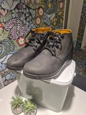 Joya Manchester svarta ankelboots, läder, storlek 42 8.5 - Säljer ett par stilrena grå kängor med svart snörning och rund tå. Skorna har en mjuk gul insida som ger en snygg kontrast. Tillverkade i skinn med platt sula, perfekta för dig som gillar enkel och clean design.