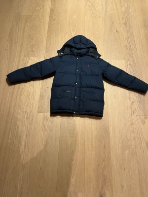 Mörkblå pufferjacka från Polo Ralph Lauren - Snygg mörkblå pufferjacka från Polo Ralph Lauren med huva och långa ärmar. Jackan har broderad röd logga på bröstet, knappar och dragkedja framtill. Perfekt för kalla dagar och har en klassisk, stilren look. Den är köpt från kindsbrandstore och säljs för att den var förliten för mig. Storleken är L barn