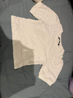 Vit blus med struktur från Vero Moda - Säljer en vit blus från Vero Moda i storlek M. Blusen har ett snyggt strukturerat tyg med diskret mönster och puffiga korta ärmar. Rund halsringning och normal passform. Perfekt för dig som gillar stilrena detaljer och mjukt material.