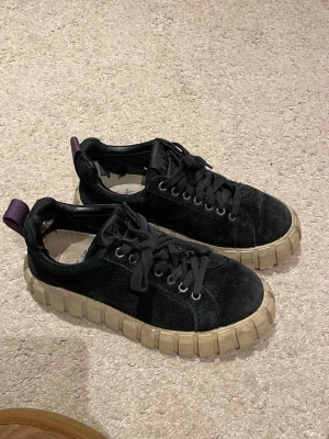 Svarta Eytys sneakers med grov sula - Säljer ett par svarta sneakers från Eytys i storlek 38. Skorna har en chunky beige gummisula och ovandel i mocka med snörning. Detalj med lila flärp bak. Perfekta för dig som gillar streetstyle och vill ha något som sticker ut.