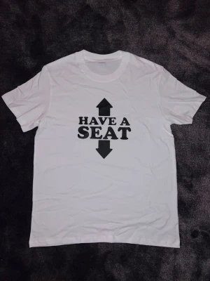 ”Have a seat” T-shirt - En vit t-shirt med ett enkelt och humoristiskt tryck. Texten “Have a seat” kompletteras av pilar uppåt och nedåt, vilket ger ett lekfullt budskap. Perfekt som rolig vardagströja eller till festliga sammanhang. Tröjan är i nytt skick och bekväm att bära.