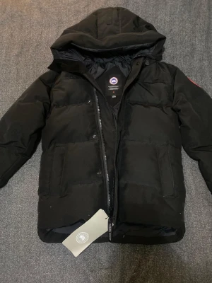 Svart pufferjacka från Canada Goose - Svart pufferjacka från Canada Goose med huva och klassisk logga på ärmen. Jackan har två stora fickor framtill och dragkedja. Materialet är polyester och dun, perfekt för kalla dagar. Snygg och stilren design som håller dig varm hela vintern.