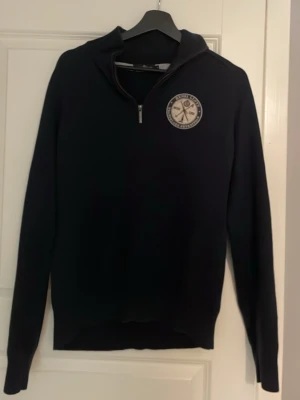 Mörkblå half zip tröja Henri Lloyd - Snygg mörkblå tröja från Henri Lloyd med half zip och broderad patch på bröstet. Tröjan har ribbad krage och långa ärmar, perfekt för dig som gillar klassisk stil med en sportig touch. Materialet är mjukt och känns skönt mot huden.