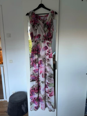 Mönstrad långklänning By Jolima - Säljer en färgstark långklänning från By Jolima med omlott-vringning och ärmlös design. Klänningen har ett unikt mönster med leopard och fjärilar i rosa, blått, grönt och brunt på en ljus bas. Materialet är lätt och luftigt, perfekt för varma dagar.