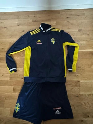 Svenskt Adidas fotbollsställ - Säljer ett mörkblått och gult fotbollsställ från Adidas med Sveriges landslagsemblem på både jacka och shorts. Jackan har dragkedja, gula detaljer och Bauhaus-logga. Perfekt för dig som gillar fotboll och vill ha landslagskänsla på träningen.