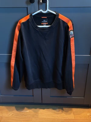 Svart fleece tröja Parajumpers S - Svart fleece tröja från Parajumpers i storlek S. Snygga orange detaljer längs ärmarna med Parajumpers-text och patch på axeln. Klassisk rund halsringning och ribbade muddar. Perfekt för dig som gillar sportig och clean stil.