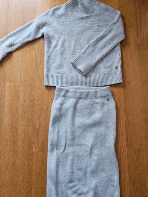 Ljusgrå ribbad stickad set - Mysig ljusgrå stickad tröja med ribbad struktur och hög hals. Tröjan har långa ärmar och en avslappnad passform, perfekt för kyliga dagar. Enkel och stilren design som passar till det mesta.