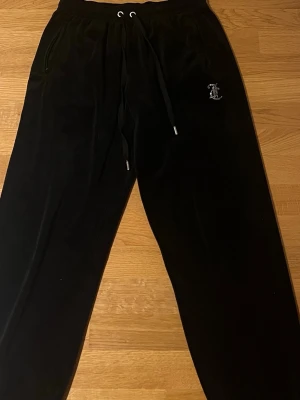 Svarta Juicy Couture - Svarta mjukisbyxor från Juicy Couture i storlek S. Byxorna har snörning i midjan, fickor och ett glittrigt Juicy-tryck på baksidan. Materialet är mjukt och sammetliknande, perfekt för chill dagar. Klassisk Juicy Couture-logga på framsidan.
