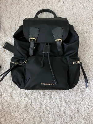 Svart Burberry ryggsäck med gulddetaljer - Säljer en snygg svart ryggsäck från Burberry med guldfärgade metalldetaljer och quiltad baksida. Väskan har flera dragkedjefickor, justerbara axelremmar med kedjedetaljer och klassisk Burberry-logga framtill. Materialet är en mix av nylon och skinn för en lyxig känsla.