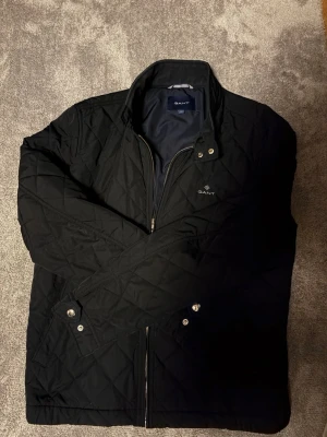 Svart quiltad jacka från GANT - Quiltad svart jacka från GANT med dragkedja och tryckknappar vid fickor och ärmslut. Klassisk krage och diskret logga på bröstet. Jackan har ett stilrent rutmönster och är perfekt för kyliga dagar. Snygg och enkel att matcha med olika outfits. 