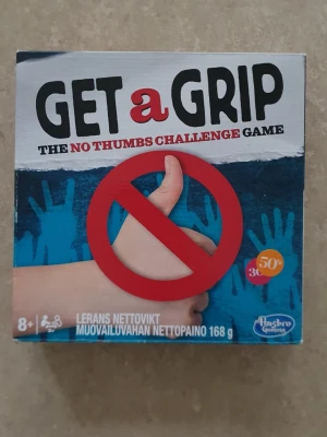 Get a Grip - The No Thumbs Challenge Game - Get a Grip är ett roligt sällskapsspel där du ska klara utmaningar utan att använda tummarna. Spelet innehåller tumhandskar med rött och vitt mönster, modellera, kort och tillbehör. Perfekt för att utmana vänner och familj på kreativa och knasiga uppgifter.