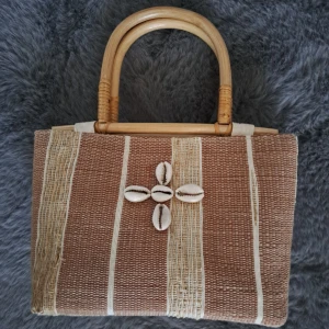 Beige handväska med snäckor och bambuhandtag - Snygg handväska i beige och brunt med naturliga bambuhandtag. Väskan har ett randigt mönster och är dekorerad med snäckor framtill. Tillverkad i vävt material och stängs med dragkedja. Perfekt accessoar för en chill sommarlook.