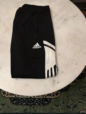 Svarta Adidas träningsbyxor - Svarta träningsbyxor från Adidas med vit logga och vita detaljer på sidan, samt klassiska ränder. Byxorna har elastisk midja och är tillverkade i polyester. Perfekta för sport och träning, med snygg och sportig design.
