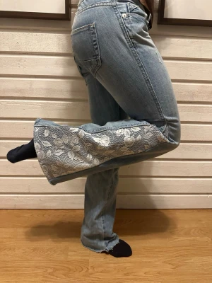 Bootcut jeans med spets från Gina Tricot - Unika ljusblå bootcut jeans från Gina Tricot med breda spetsinsatser i vitt blommönster längs benen. Jeansen har klassisk femficksdesign, råa kanter nertill och är tillverkade i bomullsmaterial. Perfekt för dig som vill sticka ut med en cool och personlig stil.