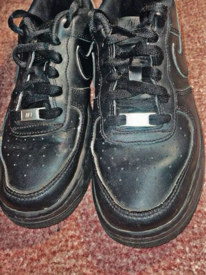 Nike Air Force 1 svarta sneakers - Svarta Nike Air Force 1 sneakers med klassisk låg siluett och rund tå. Skorna har snörning med svarta skosnören och detaljer i metall framtill. Tillverkade i skinn med perforeringar på tån för extra ventilation. Swoosh-logga på sidorna och mönstrad platt sula.