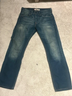 Levi's 533 Loose blå jeans - Säljer ett par Levi's 533 Loose jeans i klassisk blå tvätt med snygga slitningar och kontrastsömmar. Modellen har loose fit och raka ben, fem fickor och knappgylf. Perfekta för dig som gillar avslappnad stil och vill ha gott om rörelsefrihet.