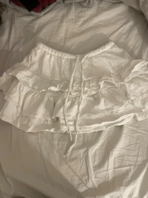 Vit volangkjol med snörning - Säljer en vit kortkjol med flera lager volanger och snörning i midjan. Kjolen har en mjuk och luftig känsla, perfekt för dig som gillar en feminin och lekfull stil. Elastisk midja gör den enkel att ta på och av. Materialet känns som bomull.