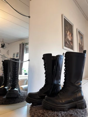 Svarta höga boots  - Säljer ett par svarta höga boots i storlek 39 med grov sula och snörning hela vägen upp. Skorna har dragkedja på insidan för enkel påtagning och är tillverkade i skinnimitation. Från märket VOX.  URSPRUNGLIGT PRIS: 600 KR