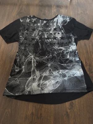 Svart t-shirt med stjärnmönster - Cool svart t-shirt med korta ärmar och ett unikt tryck av vita och gråa rökformationer samt stjärnor över bröstet. Perfekt för dig som gillar grafiska detaljer och vill sticka ut lite extra. Skön passform och rund halsringning.