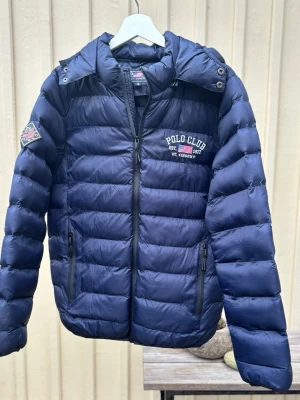 Mörkblå pufferjacka Polo Club S - Snygg mörkblå pufferjacka från Polo Club med broderad logga på bröstet och ärmen. Jackan har huva med tryck, två fickor med dragkedja och quiltad design. Perfekt för kalla dagar.