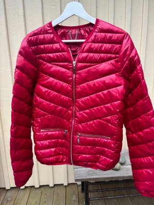 Röd pufferjacka - Snygg röd pufferjacka från Amisu med glansig finish och quiltad design. Jackan har rund hals, långa ärmar och två framfickor med dragkedja. Perfekt för dig som vill ha en lätt och cool jacka med modern look.