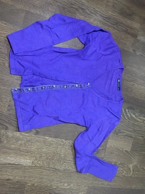 Lila kofta från Elexa med knappar - Snygg lila kofta från Elexa i storlek L/XL men mer en M. Koftan har långärmade ärmar, v-ringad hals och glittriga knappar framtill. Materialet är mjukt och stickat, ger extra lyxig känsla. Perfekt att slänga på sig när det är lite kyligt ute.