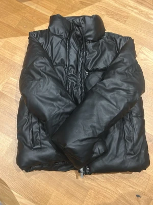 Svart pufferjacka från 157, strl 140 - Svart pufferjacka från 157 med hög krage och dragkedja. Yttermaterialet är i syntet (PU) och fodret är polyester, vilket gör jackan både varm och mjuk. Perfekt för kalla dagar och har en clean look som funkar till streetwear. Snygg och enkel design.