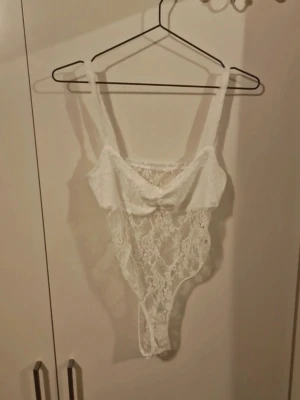 Vit spetsig korsett-topp - Säljer en vit korsett-topp i spets med smala axelband och genombrutet mönster. Toppen har en feminin och romantisk look med dekorativ rosett framtill. Perfekt för att skapa en snygg layering eller som statement-plagg.