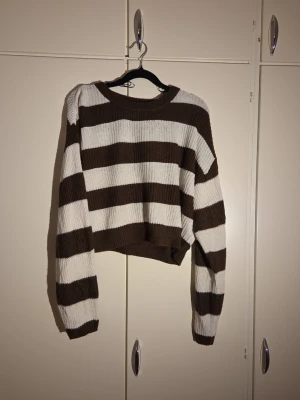 Randig brun och vit stickad tröja H&M - Säljer en stickad tröja från H&M Divided i storlek S. Tröjan har breda bruna och vita ränder, rund hals och är croppad i modellen. Materialet är 100% akryl och den har långa ärmar. Perfekt för dig som gillar en chill och trendig look.