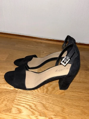 Svarta klackskor Säljes! - Svarta klackskor från Deichman med bred rem över tårna och justerbart spänne runt ankeln.  I storlek 39