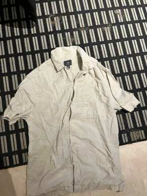 Beige kortärmad skjorta från H&M - Säljer en stilren beige skjorta från H&M i luftig linnemix. Skjortan har korta ärmar, klassisk krage, bröstficka och knäppning framtill. Perfekt för varma dagar och enkel att matcha med olika outfits. Säljer pga inte känner användning utav den i vinter.