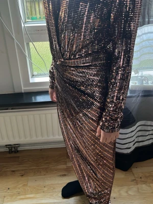 Glittrig långklänning med slits - Säljer en glittrig långklänning i svart och guld med snygga vertikala ränder. Klänningen har lång ärm, omlott-design och en djup slits framtill. Materialet är syntet med paljetter som ger en riktigt festlig look. Perfekt för dig som vill sticka ut och glänsa.