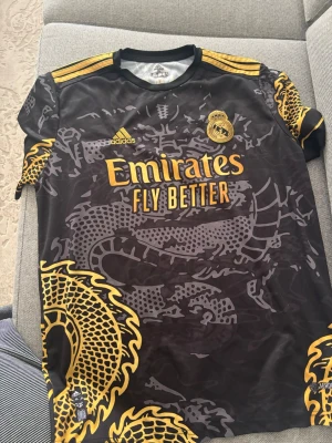Real Madrid x Adidas Ronaldo tröja XL - Säljer en Real Madrid matchtröja från Adidas i storlek XL med Ronaldo och nummer 7 på ryggen. Tröjan är svart med guldiga detaljer och drakmönster, samt gula Adidas-ränder på axlarna. Materialet är Aeroready polyester som andas.