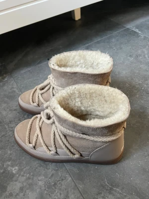 Beige Inuikii boots med snörning - Säljer ett par beige Inuikii boots med grov snörning och mjukt, fluffigt foder. Skorna har en rund tå, platt sula och är tillverkade i mocka och skinn. Perfekta för kalla dagar med sin varma insida och snygga design.