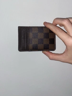 Luis Vuitton plånbok - Säljer en snygg och stilren plånbok i klassiskt brunt och mörkbrunt rutigt mönster. Plånboken är tillverkad i skinn och har flera fack för kort. Perfekt för dig som vill ha något lyxigt och praktiskt i fickan.
