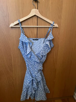 Blå omlottklänning med hjärtan från SHEIN - Supersöt blå kortklänning från SHEIN med vita hjärtan som mönster. Klänningen har omlottmodell, volangdetaljer och smala axelband. Knytband i midjan för extra passform. Perfekt för dig som gillar romantisk och lekfull stil.