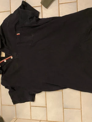 Svart henley från FILR med kort ärm - Svart henley med korta ärmar från FILR. Skjortan har vit- och röd-detaljer vid knappraden och en bröstficka med liten röd-vit etikett. Tillverkad i mjuk bomull, perfekt för chill dagar. Klassisk passform och stilren look.