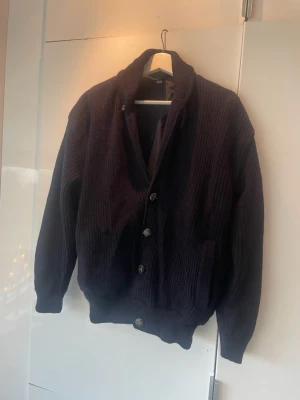 Mörkblå stickad kofta med knappar - Säljer en mörkblå, grovstickad kofta med snygga knappar framtill och ribbade muddar. Koftan har en skön, lite loose passform och är tillverkad i ren ny ull och bomull. Perfekt för kyliga dagar och enkel att matcha med jeans eller chinos. Den är m men sitter som s