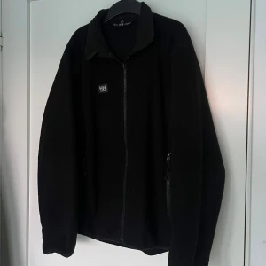 Svart fleecekofta från Helly Hansen - Svart fleecekofta från Helly Hansen med hel dragkedja framtill och hög krage. Tröjan har två sidofickor med dragkedja och HH-logga på bröstet. Perfekt för kyliga dagar och enkel att slänga på över en t-shirt.