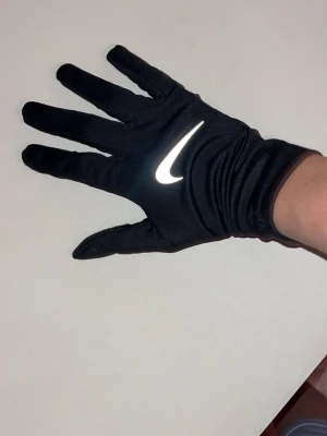 Svart Nike thermal fit  träningshandskar - Svarta träningshandskar från Nike med reflekterande logga på ovansidan. Handskarna är tunna och smidiga, tillverkade i syntetmaterial med förstärkt tumme för extra grepp. Perfekta för löpning eller gym. Snygg och sportig design som passar dig som gillar att röra på dig.