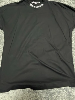 Svart Palm Angels t-shirt med tryck - Svart t-shirt från Palm Angels med stort vitt tryck på ryggen och mindre tryck vid halsen. Klassisk rund hals och korta ärmar. Materialet känns mjukt och lätt, perfekt för en chill streetwear-look. Passformen är loose och t-shirten har en enkel, stilren design.