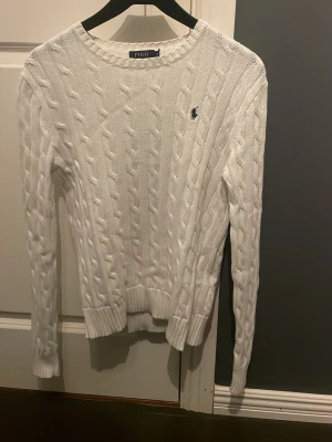 Vit kabelstickad tröja Polo Ralph Lauren - Kabelstickad vit tröja från Polo Ralph Lauren med rund halsringning och diskret logga på bröstet. Långärmad modell i mjuk bomull som passar perfekt till jeans. Klassisk och tidlös design med ribbade muddar.