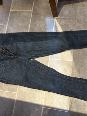 Mörkblå jeans från Emilio, storlek 28 - Snygga mörkblå jeans från Emilio med klassisk femficksdesign och röda sömmar som ger en cool detalj. Tillverkade i 99% bomull och 1% lycra för lite stretch och skön passform. Perfekta för dig som gillar stilrena och tidlösa jeans.