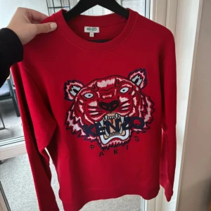 Röd Kenzo sweatshirt med tigertryck - Säljer en röd sweatshirt från Kenzo med ikoniskt broderat tigerhuvud och texten 'Kenzo Paris' framtill. Tröjan har rund halsringning och långa ärmar. 