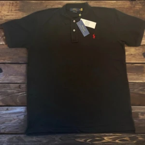 Svart pikétröja från Polo Ralph Lauren - Svart pikétröja från Polo Ralph Lauren med klassisk krage, korta ärmar och knappar framtill. Tröjan har den ikoniska röda Polo-loggan broderad på bröstet. Perfekt för dig som gillar stilrena och tidlösa plagg.