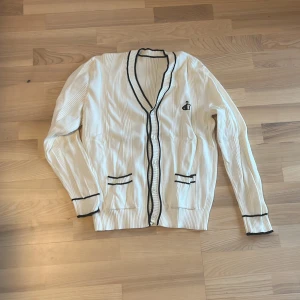 Vit kofta med svart broderad logga - Snygg vit med svarta detaljer och broderad logga på bröstet. Koftan har ribbad struktur, knappar framtill, två fickor och svarta kantband vid ärmslut, fickor och hals.💕💕