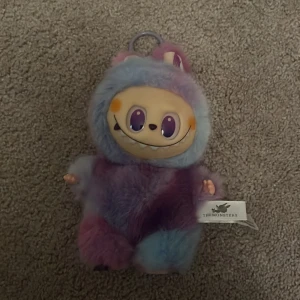 Färgglad Labubu  TheMonsters - Labubu med ett gulligt monsteransikte med stora lila ögon och ett brett leende. Perfekt som statement piece för dig som gillar roliga accessoarer.